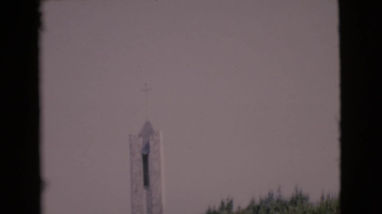 Distant white church steeple above trees under a pale sky, framed by dark film edges.