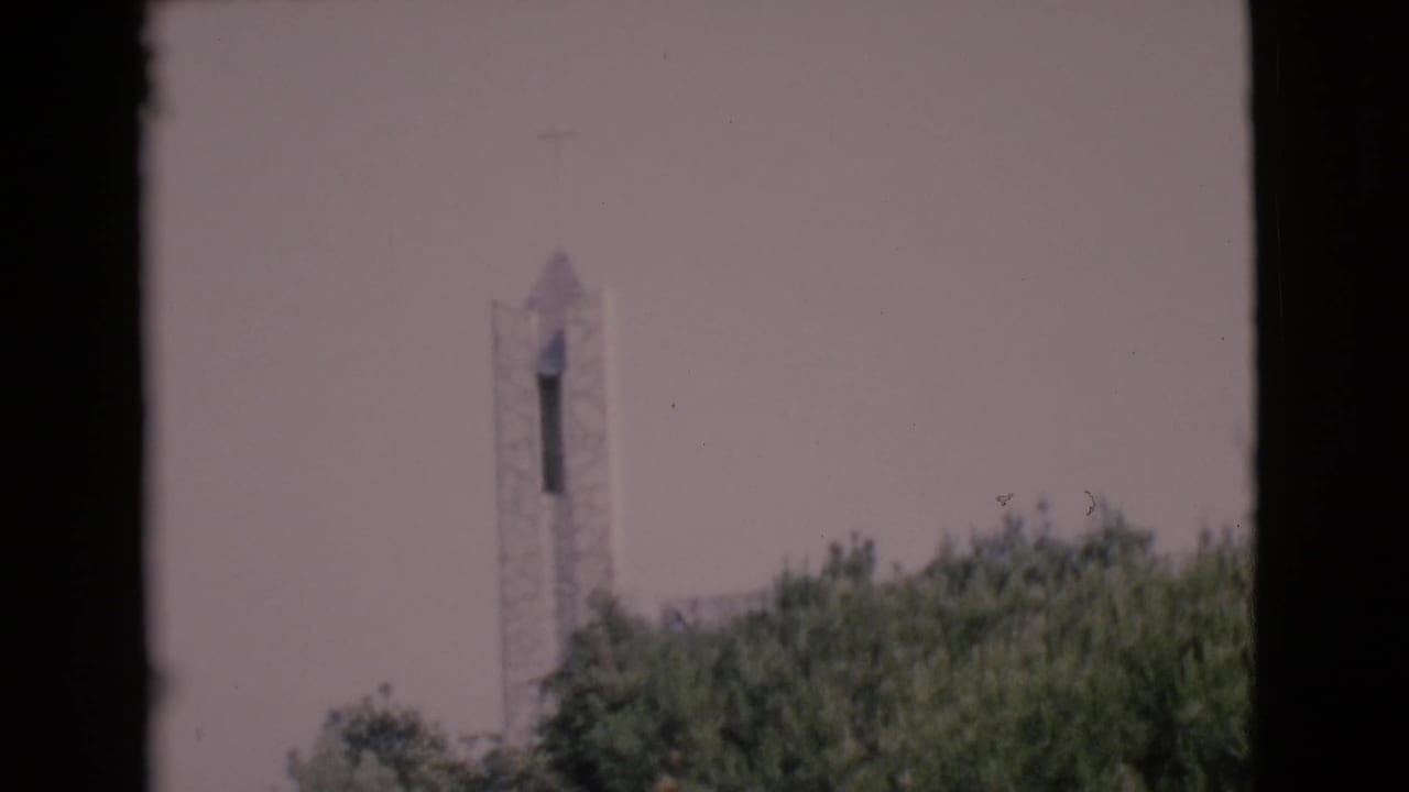 1968 8mm frame showing a distant chapel tower and cross above green treetops, likely in Southern California.