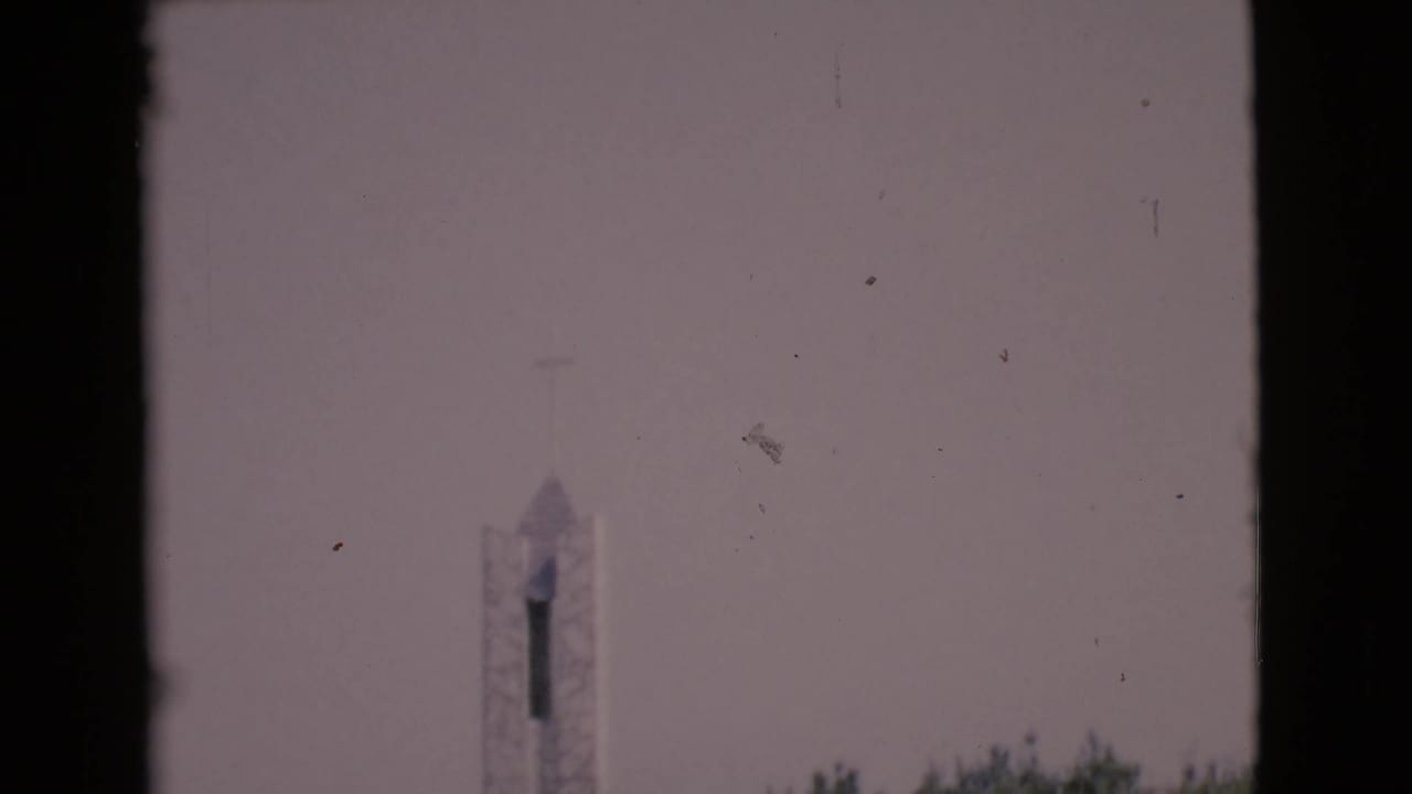 A distant stone chapel tower with a cross is seen against a pale sky in soft 1968 home-movie footage.