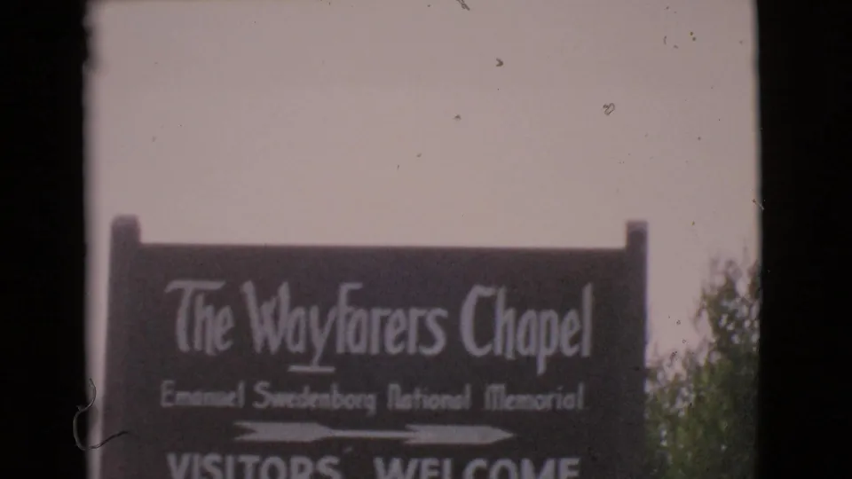 1968 8mm frame showing a dark roadside sign for The Wayfarers Chapel with white lettering and trees at the edge.