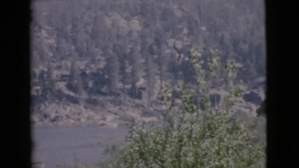 1968 San Diego County 8mm frame showing a lake, rocky opposite shore, dense trees, and dark window edges in the foreground.