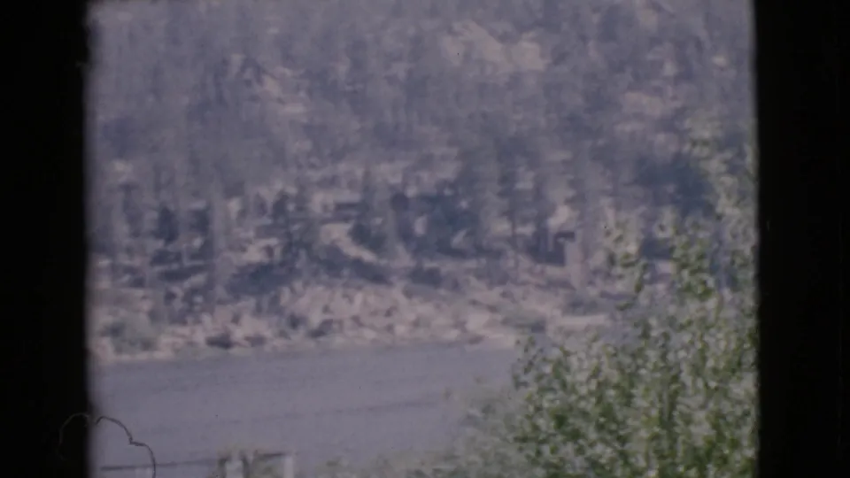1968 San Diego County home-movie frame with calm water, forested shoreline, rocks, and a dark window divider through the scene.