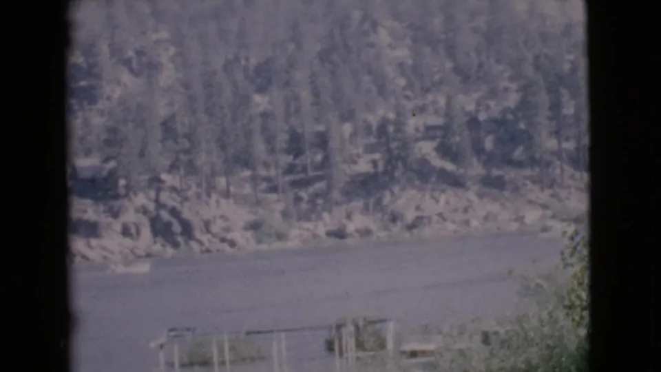 1968 San Diego County 8mm still showing calm lake water below a wooded rocky shoreline, with nearby brush at the lower edge.