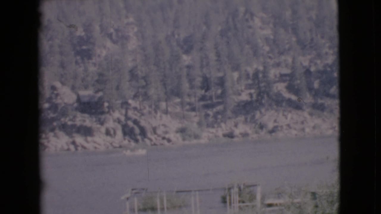 1968 San Diego County 8mm frame showing small boats on still lake water with a tree-covered rocky shore behind them.