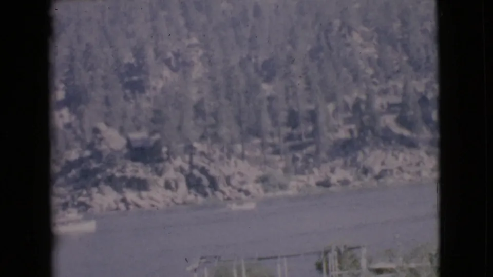 Faded 1968 8mm frame from San Diego County showing lake water, small boats, and a tree-covered rocky shoreline.