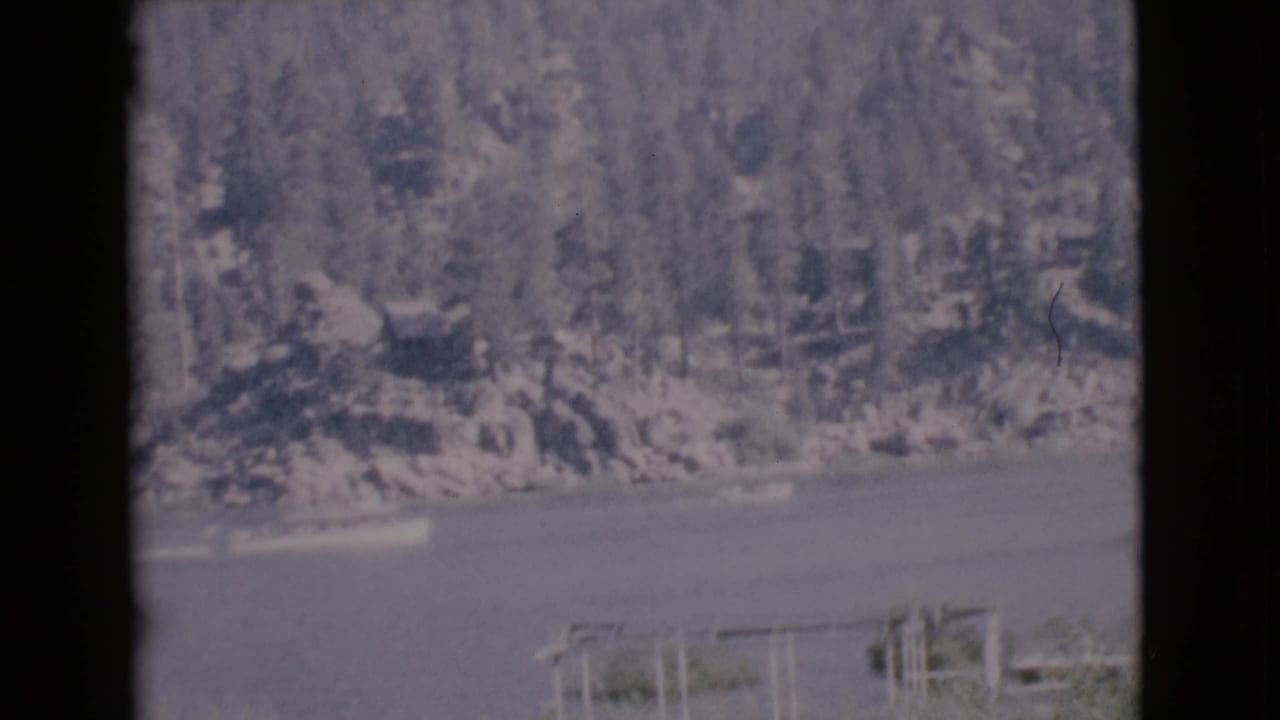 Small boat on calm lake below a forested shoreline with dark film-frame borders.