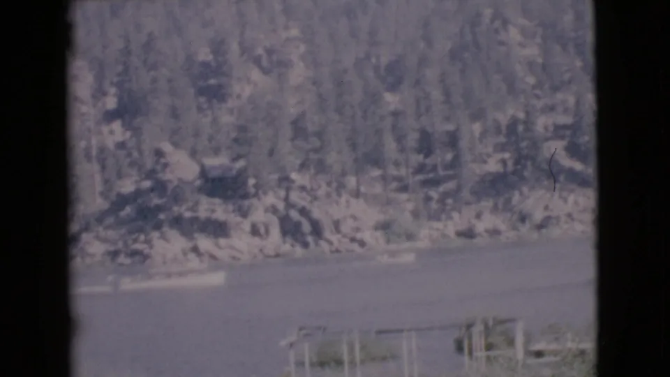 1968 San Diego County 8mm frame showing a small boat crossing calm lake water beneath a wooded, rocky shoreline.