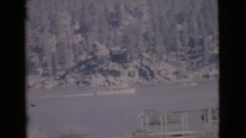1968 San Diego County lake view with a small boat near the wooded shore and a dock or railing partly visible in the foreground.