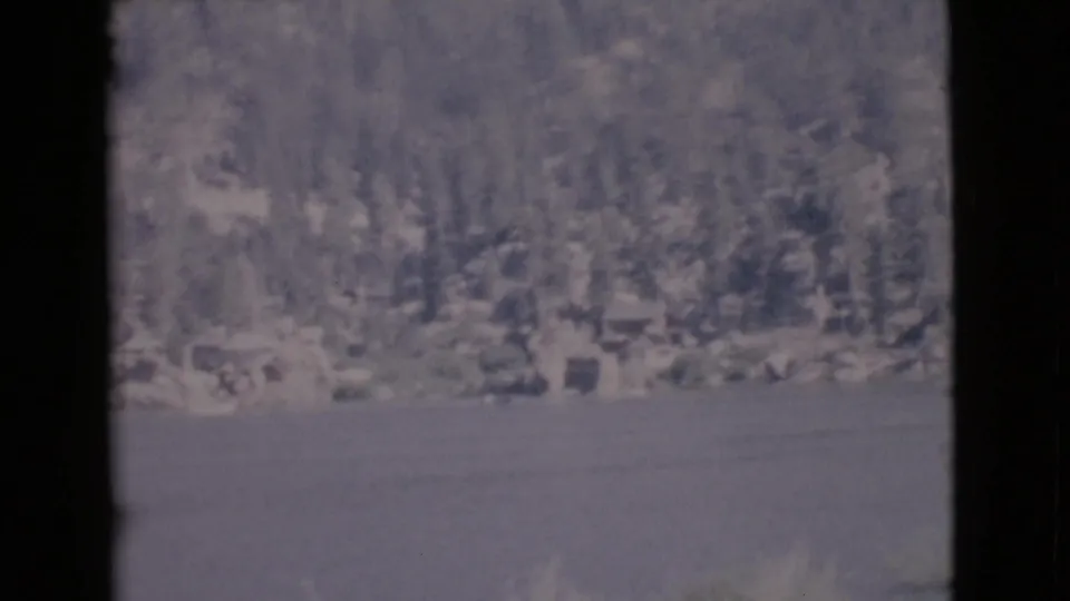 1968 home-movie frame showing calm lake water, a forested hillside, rocky shoreline, and a dock or railing at the edge of view.