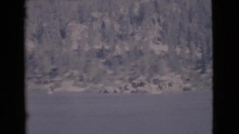 Distant lake view with wooded slope and shoreline, seen through dark 8mm frame edges in 1968 home-movie footage.