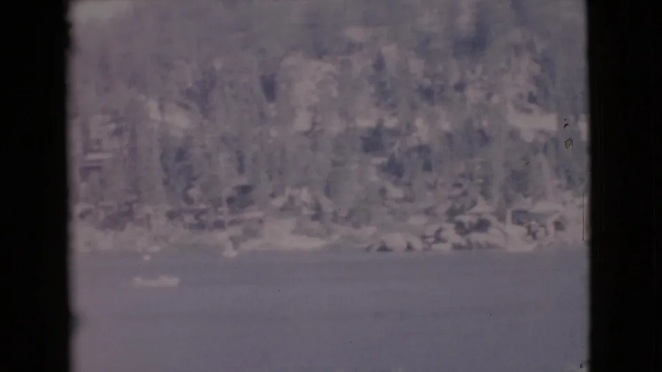 Soft 8mm lake scene showing water, a wooded shore, and faint small boats or white shapes near the shoreline.