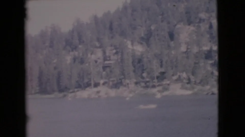 Late-1960s 8mm frame of a calm lake with wooded shoreline and small bright boat-like shapes on the water.