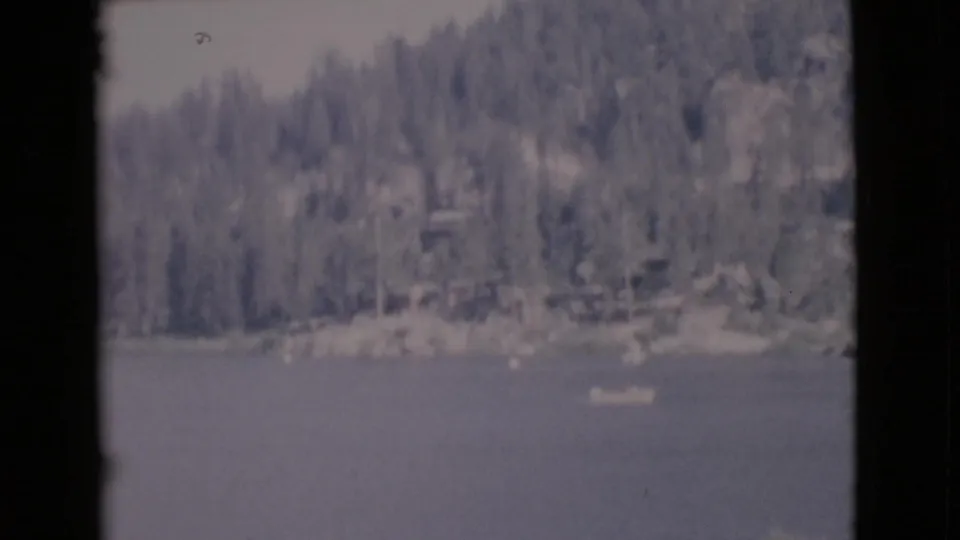 1968 8mm view of a small boat on a San Diego County lake with evergreen trees and rocky shoreline in the distance.