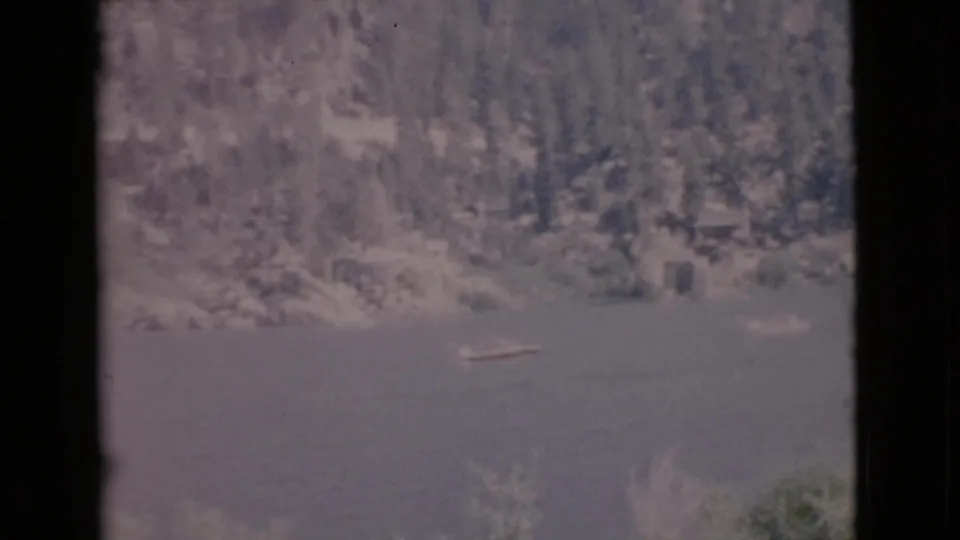 Vintage 8mm frame of a small boat crossing calm lake water beneath a forested San Diego County shoreline.