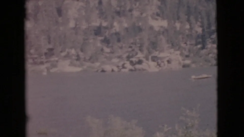 1968 San Diego County lake view with a small boat on the water, wooded shore beyond, and reeds near the lower edge.