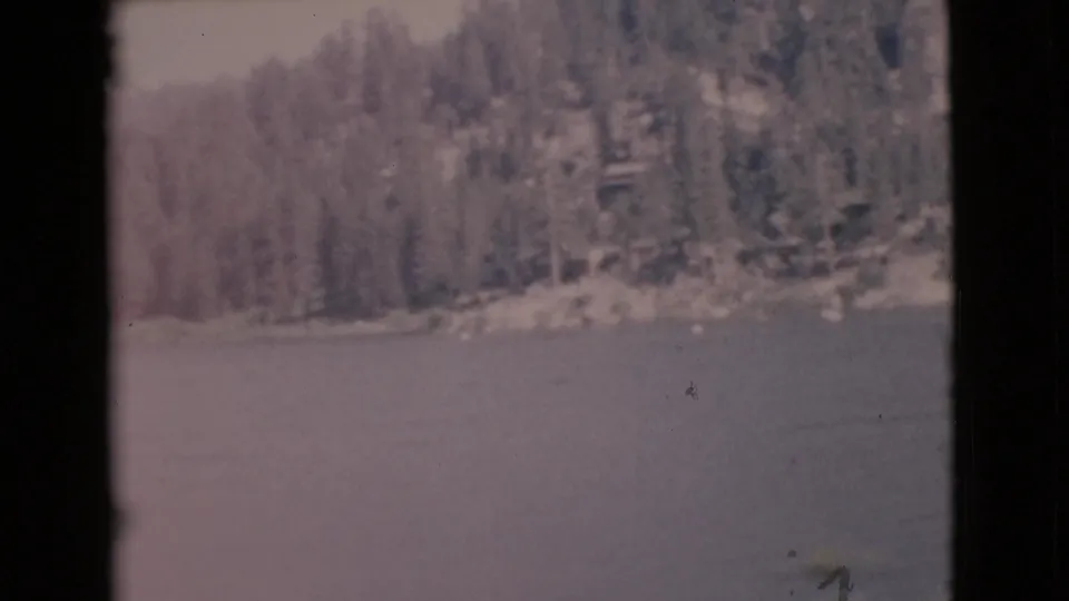 1968 home-movie frame showing lake water below a wooded hillside, with faded color and soft 8mm texture.