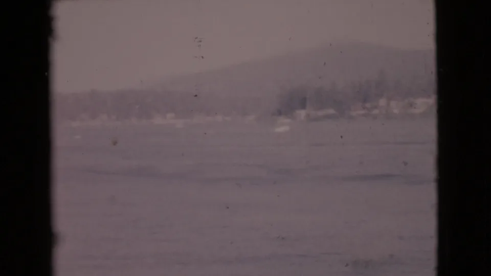 1968 San Diego County frame with calm coastal water, hazy trees and hills, faint shore structures, and visible film wear.