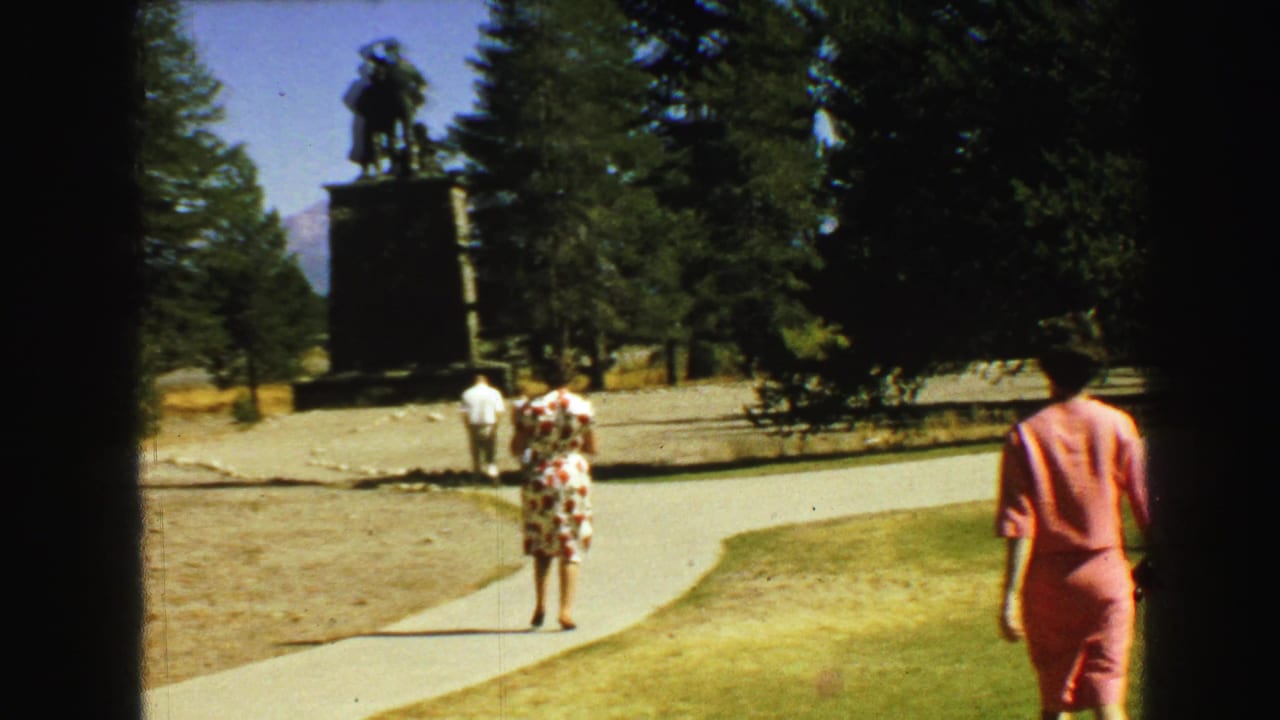 In a Vancouver park setting, adults walk along a paved path toward a tall bronze statue framed by evergreens and sunlit grass.