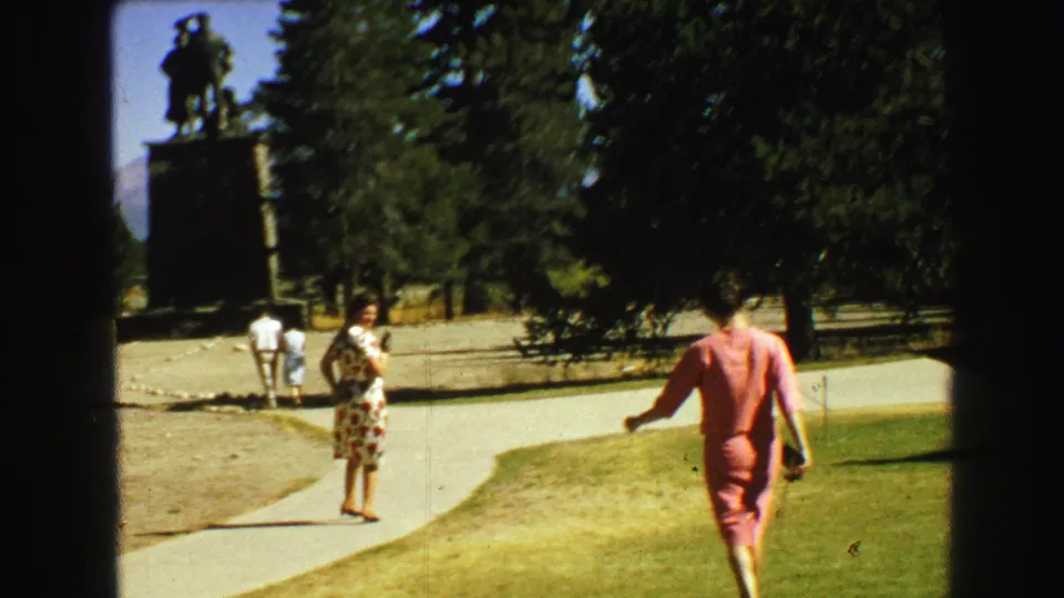 1968 home-movie footage shows adult visitors near a tall statue, including women in floral and pink dresses on a curving park path.