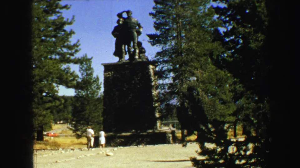 A tall bronze statue on a stone plinth stands in a Vancouver park, with adult visitors below and evergreens filling the background.