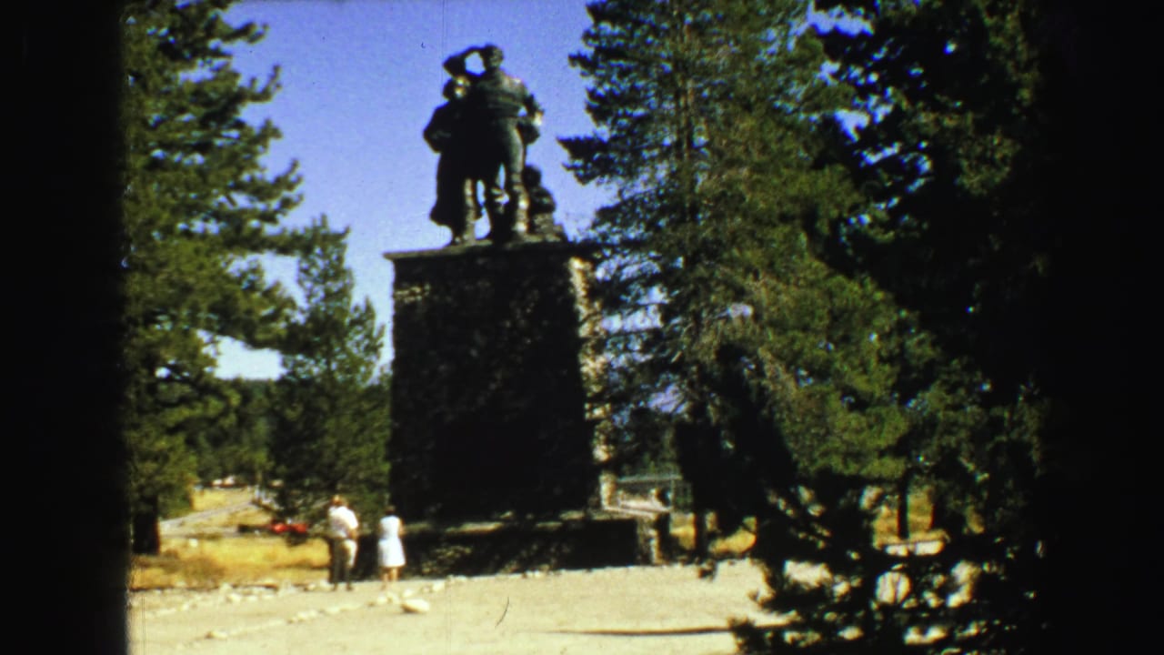 Bronze statue on a stone pedestal among pine trees with two visitors nearby.