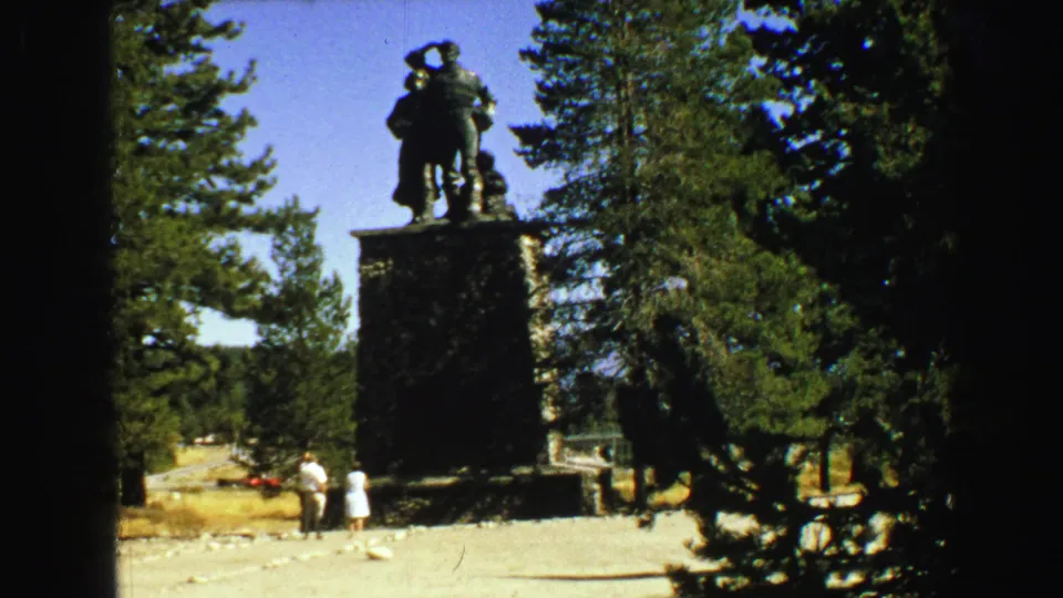 1968 Vancouver-area 8mm frame showing a large statue on a stone pedestal in a sunny forest clearing with two visitors below.