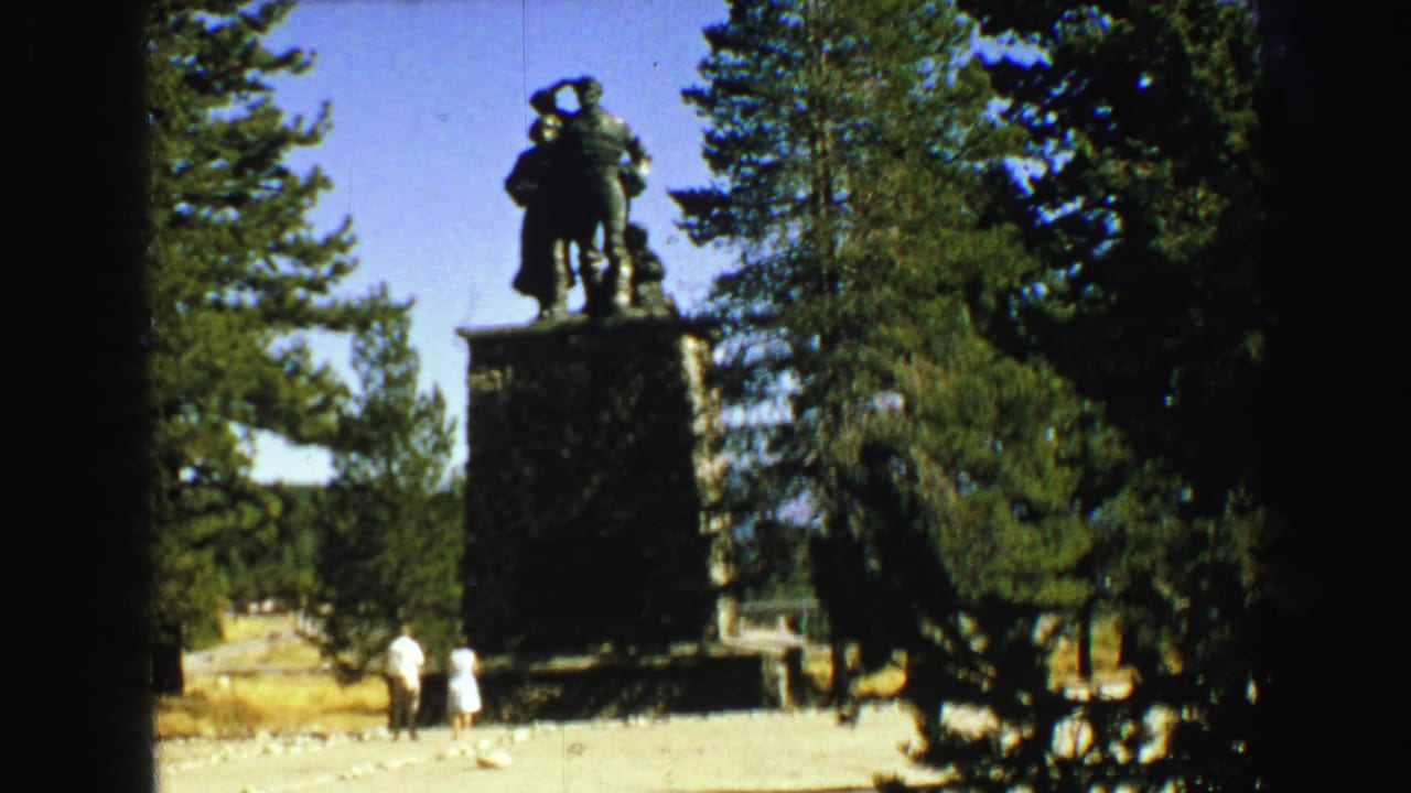 Distant visitors stand below a tall statue framed by trees.