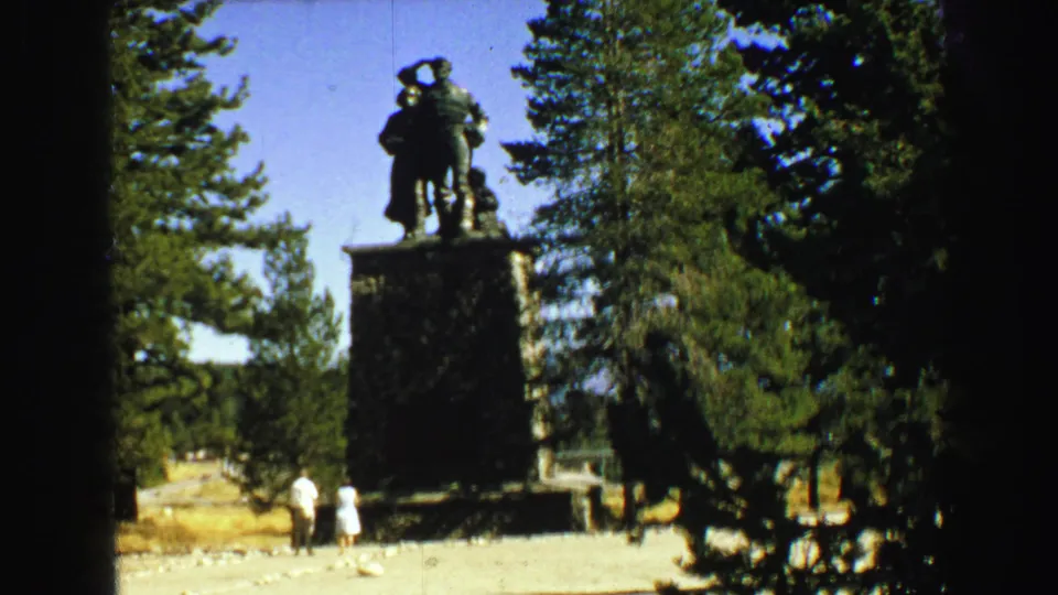 The statue and pedestal remain centered in a bright wooded setting, with two small visitor figures visible near the base.