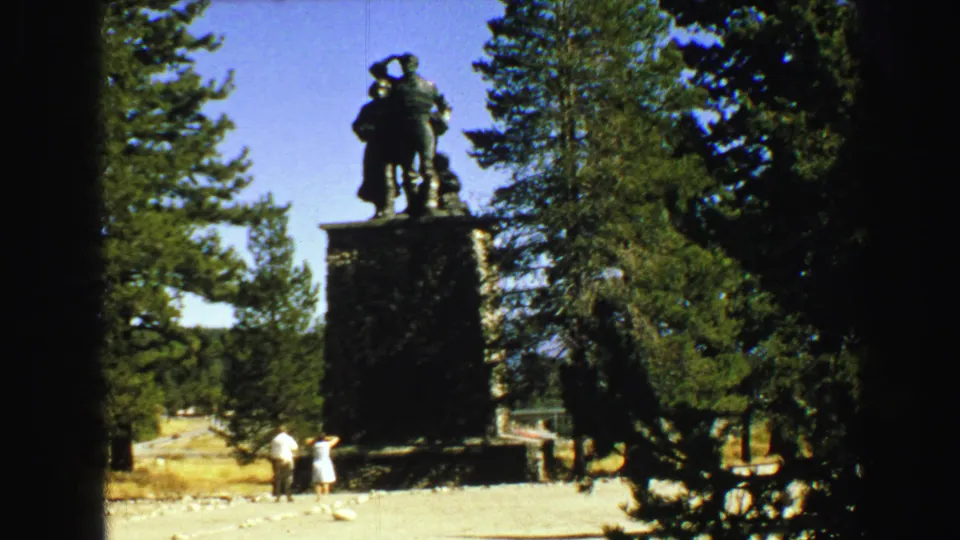 8mm frame of a stone-pedestal statue in a Vancouver forest setting, with two visitors in light period clothing below.