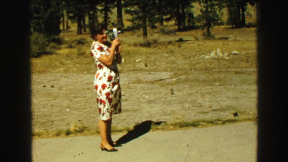 A woman in a red and white floral dress stands in the clearing holding a still camera, with trees and dry ground behind her.