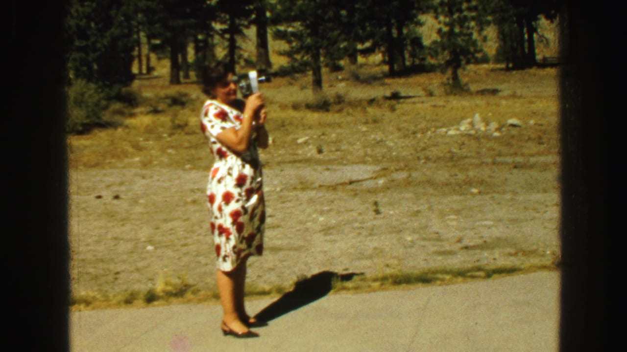 Woman in a floral dress filming with a small movie camera on a paved path.