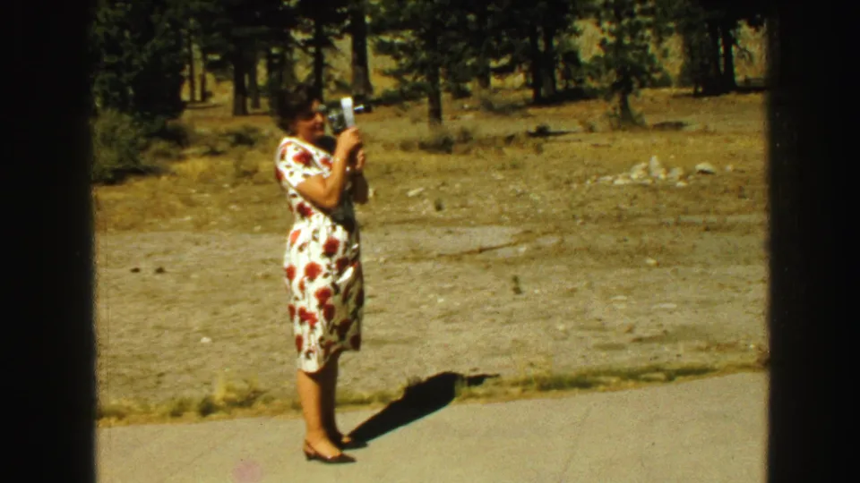 In 1968 Vancouver footage, a woman in a white floral dress films with a small 8mm camera beside a wooded path.
