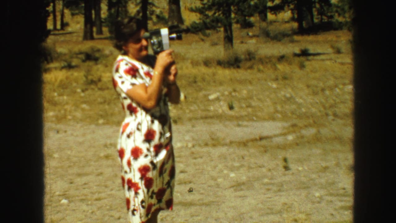 Closer view of the woman holding a small movie camera in a wooded area.