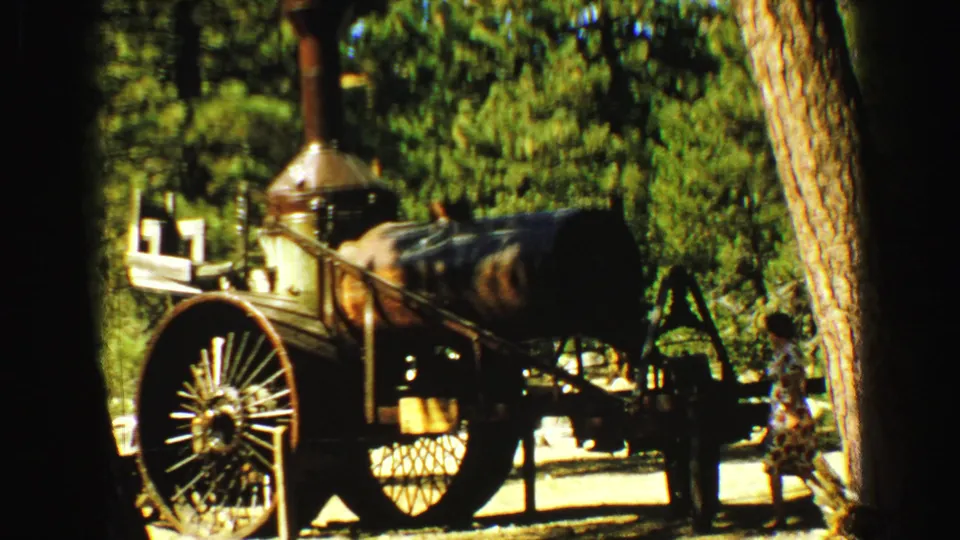 A woman in a patterned dress stands near the displayed steam engine in wooded 1968 Vancouver home-movie footage.