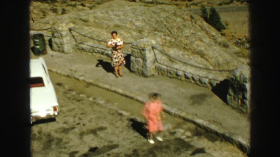 A woman in a patterned dress stands near a stone wall and chain barrier as a white classic car passes below in Vancouver.