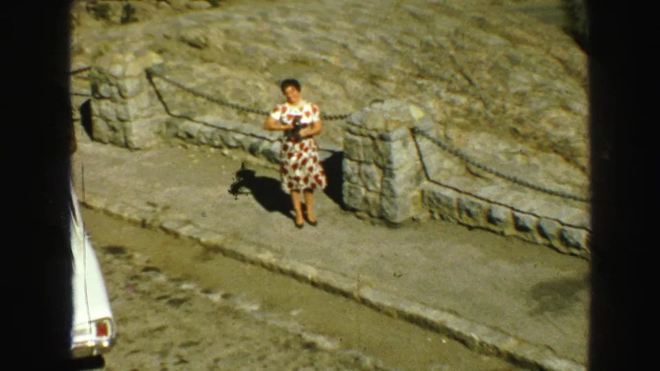 A woman in a patterned dress stands at a stone overlook with a chain barrier in 1968 Vancouver home-movie footage.