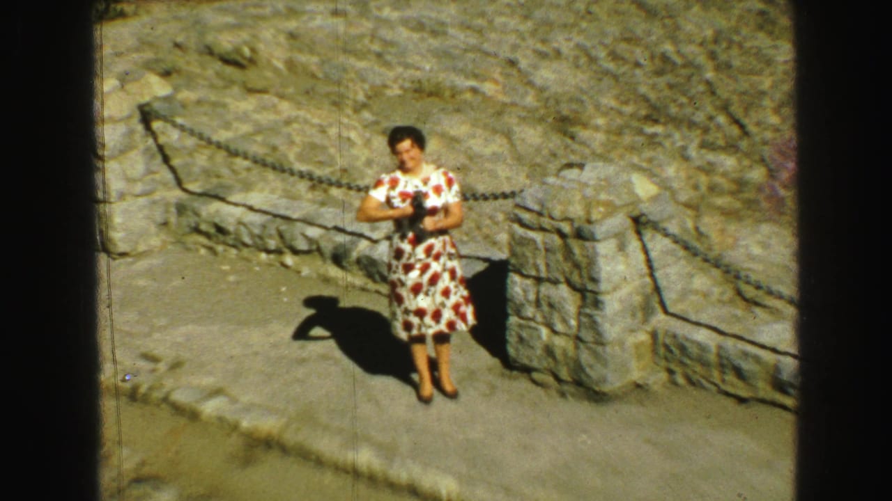Woman in a floral dress stands near a stone post and chain barrier on sunlit rock.