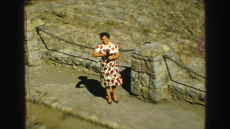 In 1968 Vancouver footage, a woman in a red and white floral dress stands with a camera beside stonework and a chain barrier.