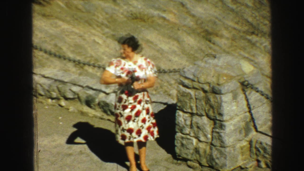 Woman in a floral dress looks aside while holding a camera near stonework and rocks.