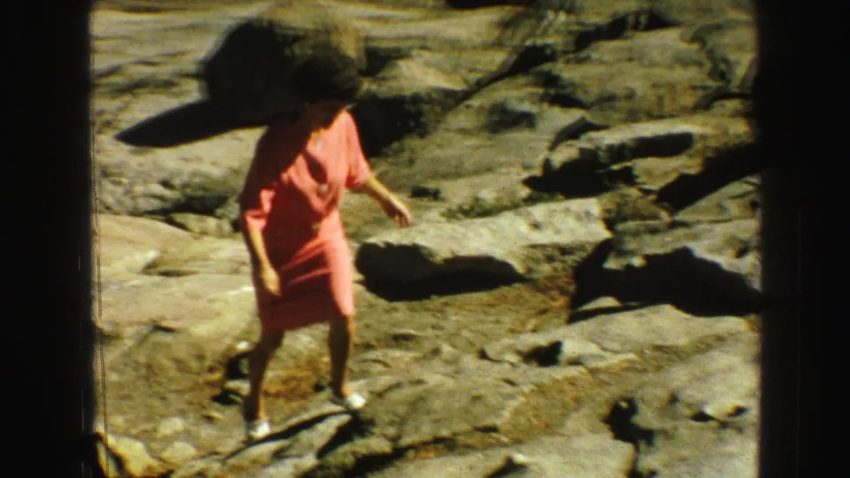 A woman in a coral dress walks carefully over uneven rocky terrain in the 1968 Vancouver home-movie footage.