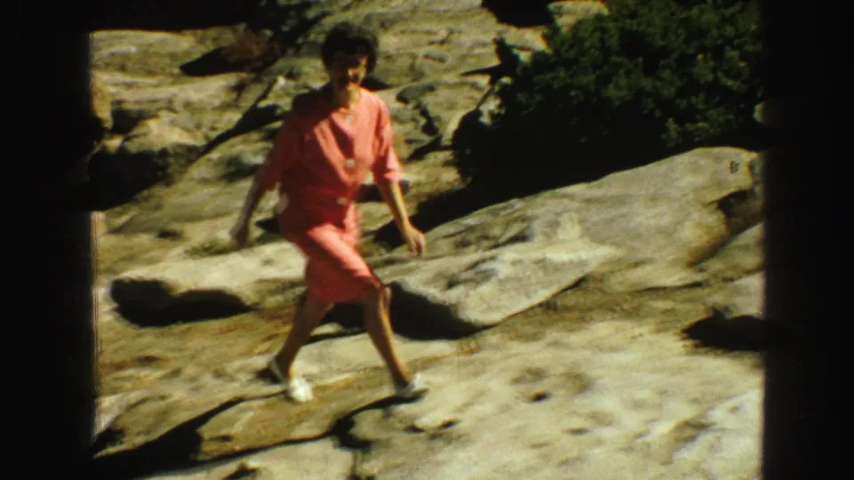 A woman in a coral dress moves across a rocky outdoor area, with shrubbery and strong shadows visible in the Vancouver footage.