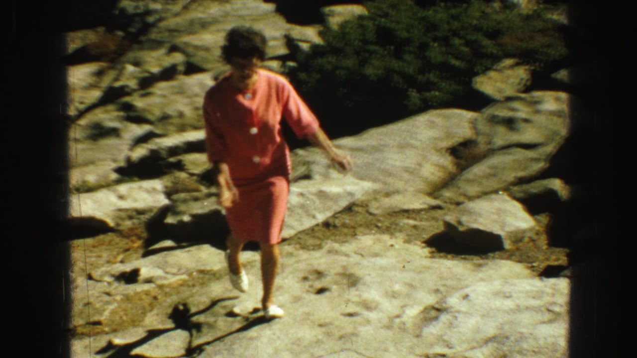 In 1968 Vancouver-area home-movie footage, a woman in coral clothing walks carefully over rocky terrain near forest.