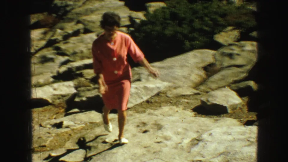 In 1968 Vancouver-area home-movie footage, a woman in coral clothing walks carefully over rocky terrain near forest.