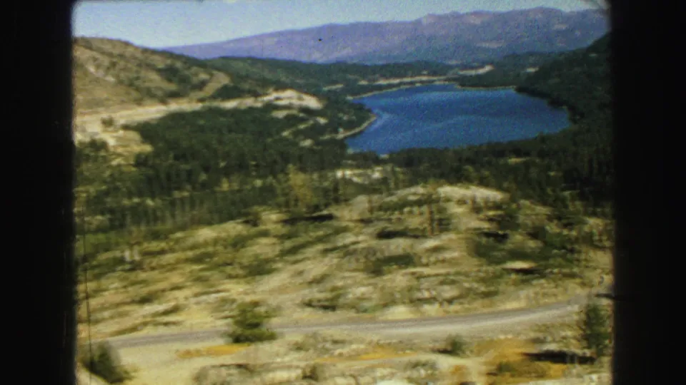 The home-movie camera frames a broad Vancouver-area mountain-lake landscape with forested slopes and rocky foreground.