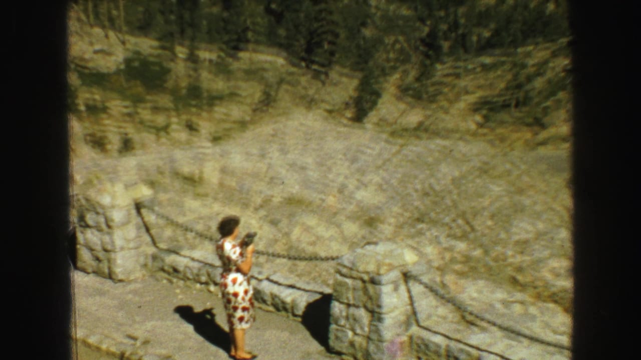 1968 Vancouver-area travel frame with a woman in a floral dress holding a camera at a stone overlook with rocky terrain behind her.