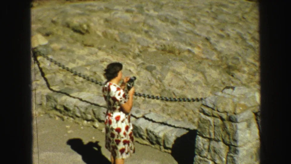 Closer 1968 home-movie view of the woman photographing from a Vancouver-area mountain overlook beside a stone post and chain railing.