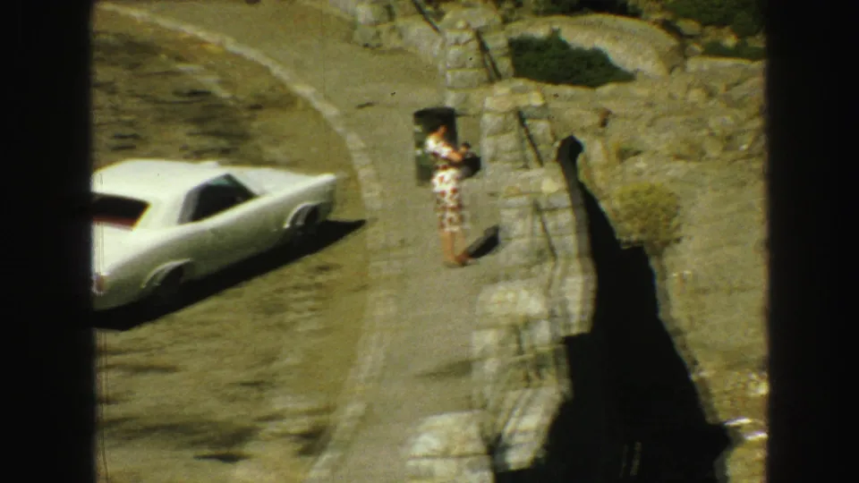1968 Vancouver-area frame showing the woman by the overlook railing near a parked white classic car on a curving road.