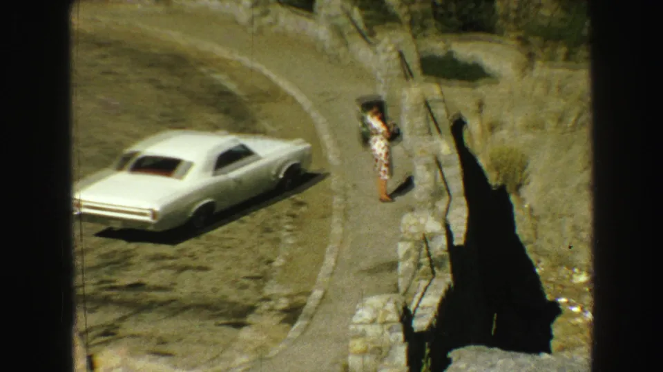 1968 Vancouver-area 8mm frame with a white car on a winding mountain road and a person standing near a rocky roadside overlook.