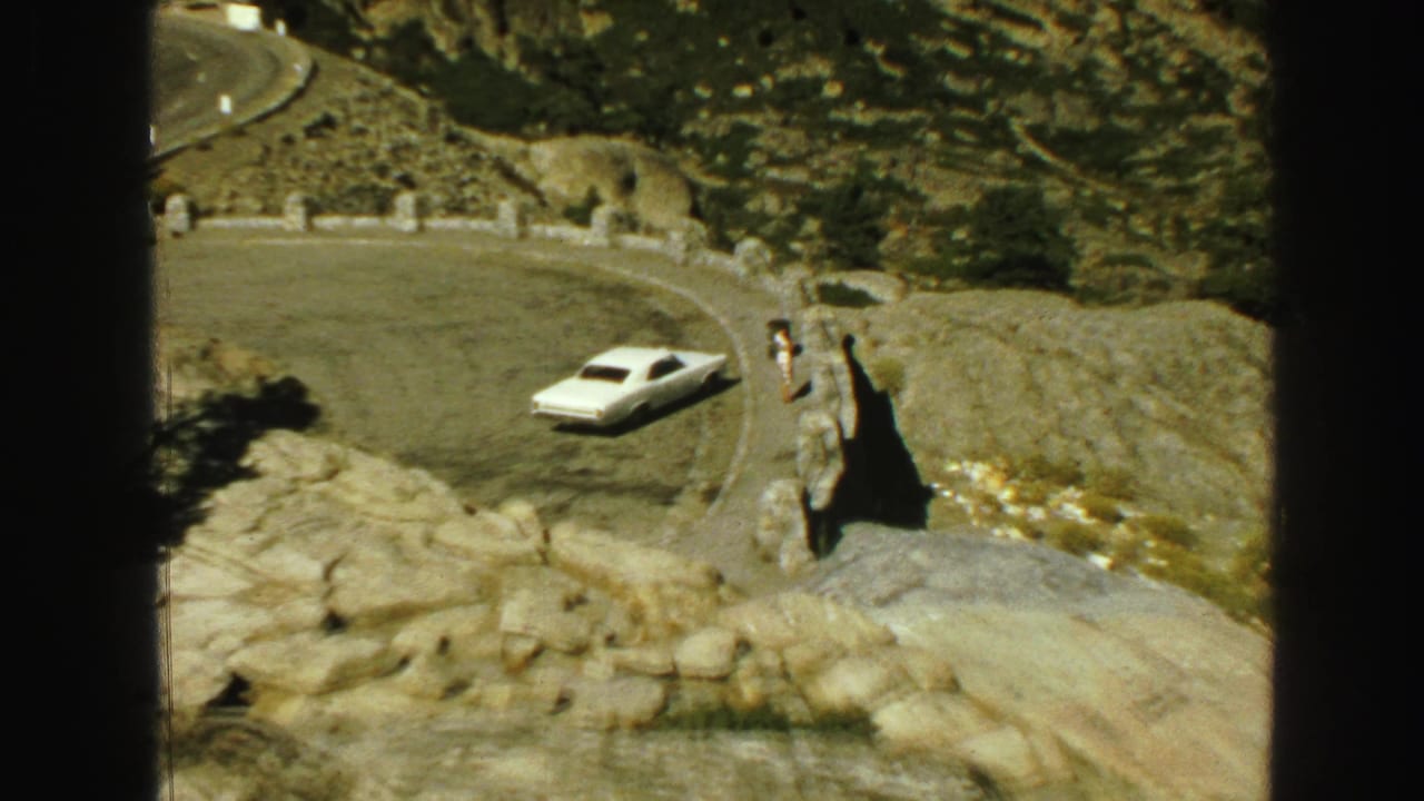 White car on a broad curve with rocky ground and roadside stones.