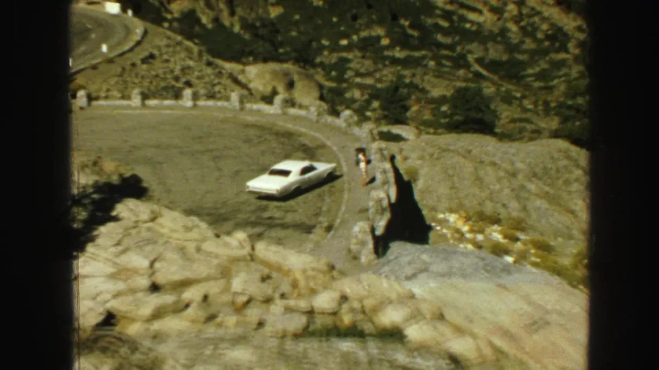 1968 Vancouver-area home-movie frame showing a white car on a mountain road curve with rocky terrain and a distant roadside overlook.